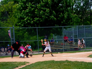 Youth baseball player preparing to swing during a summer game while teammates, coaches, and spectators watch from behind the backstop at a local baseball field in Omaha, Nebraska.