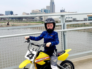Kid wearing helmet and safety gear riding an e-bike style dirt bike with Omaha skyline in background